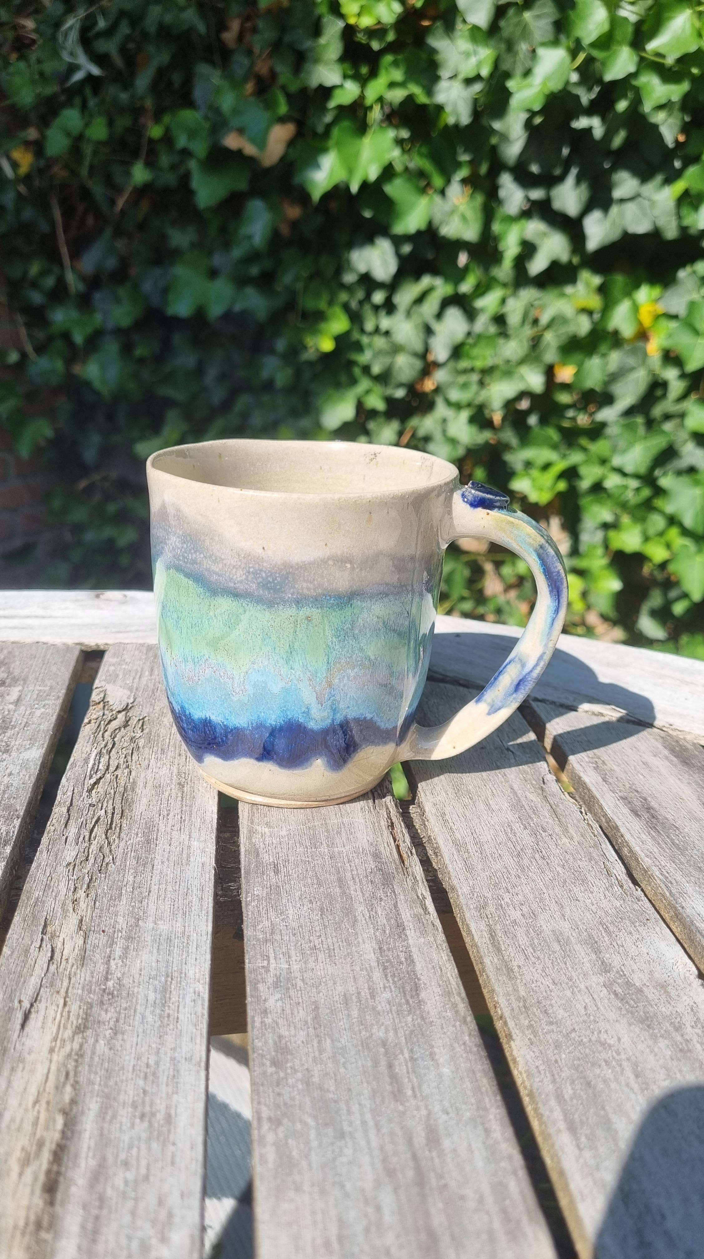 Handgemaakte keramische mok door Mira met blauw-groen en beige glazuur en handgevormde oor buiten